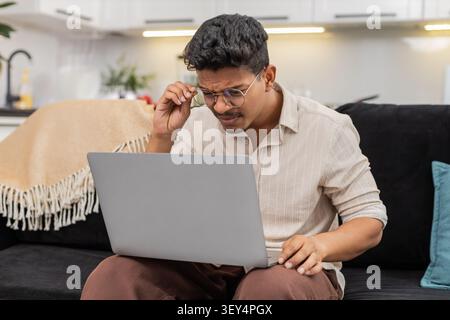 Ein indischer Mann zu Hause hat Schwierigkeiten, winzige Texte auf dem Laptop zu lesen, zieht dann eine Brille auf und sieht klare Sicht. Arabian Guy on Sofa Lächeln passt den Rahmen an, liest Bildschirmlinien ohne Belastung und setzt die Arbeit fort. Stockfoto