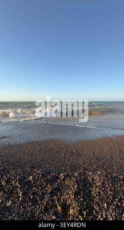 Strandwelle stürzt an Land, in Küstenszene mit klarem Himmel und fernen Bergen, mit Wellen, Sand, Steinen, mit Sand, Steinstruktur, von einem niedrigen Stockfoto