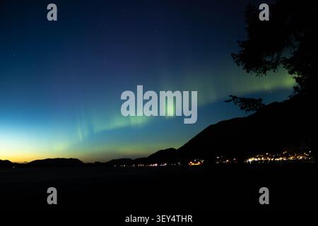 Nordlichter leuchten in der Dämmerung über einer Bergstadt an der Küste, mit Sternen, die am Nachthimmel zu sehen sind, und mit Silhouetten versehenen Bergen, Bäumen und Küsten Stockfoto