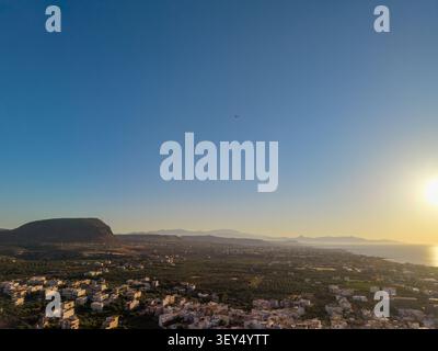 Panoramablick auf ein riesiges Küstental mit Wohnhäusern und landwirtschaftlichen Feldern in der Nähe eines Bergrückens unter Sonnenuntergang. Stockfoto