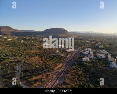 Der Aussichtspunkt der Drohne fängt eine ländliche Stadt ein, eingebettet zwischen üppigen grünen Olivenplantagen und einer zerklüfteten Bergkette unter einem klaren blauen Himmel. Stockfoto