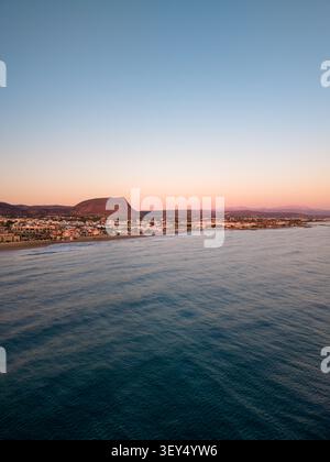 Die Hochwinkelperspektive fängt eine Küstenstadt entlang des blauen Meeres mit einem markanten flachen Berg unter einem sich abzeichnenden Sonnenuntergang-Himmel ein. Stockfoto
