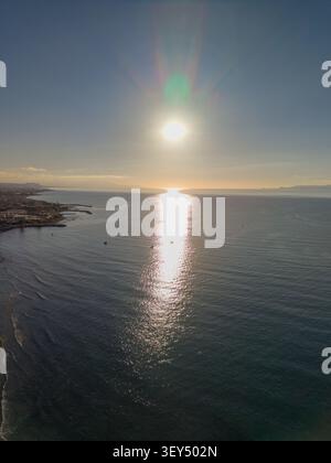 Die Hochwinkelperspektive fängt einen hellen Sonnenuntergang über der Meeresoberfläche ein und erzeugt eine starke goldene Lichtreflexion in der Nähe einer Küstenlinie. Stockfoto