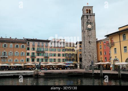 Cafés und Pastellgebäude am Wasser umgeben den Torre Apponale Uhrenturm in Riva del Garda, Gardasee, Italien, vom Hafen aus gesehen Stockfoto