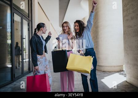 Drei Freunde kaufen zusammen, lächeln und feiern mit bunten Taschen vor einem modernen Geschäft Stockfoto