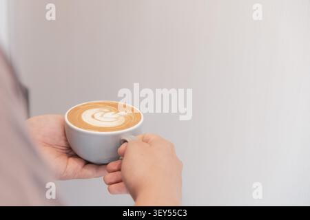 Eine einzelne Hand streckt eine graue Keramiktasse mit Latte und geschwungener Milchschaumkunst vom linken Bildrand aus, während die rechte Seite mit sauberem Weißraum ausgefüllt ist. Stockfoto