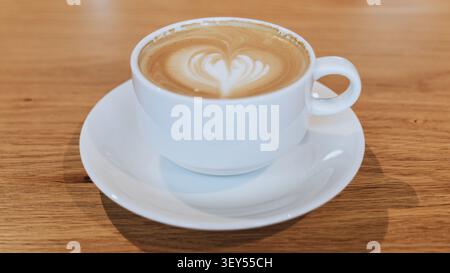 Eine weiße Keramikkaffeetasse mit Untertasse und herzförmigem Latte-Art-Design steht auf einem hellen Holztisch in einem Café. Stockfoto