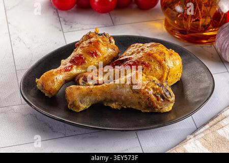 Drei goldgeröstete Hähnchenkegel auf mattschwarzer Platte. Hintergrund mit Kirschtomaten, Knoblauchzwiebeln, Glasgefäß mit Kräutern. Warme Beleuchtung Hi Stockfoto