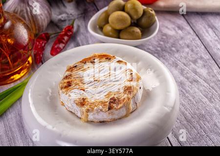 Geschmolzener Camembert-Käse auf weißer Platte mit goldener Kruste, umgeben von grünen Oliven, roten Chilischoten, Knoblauch und Olivenöl. Warme Beleuchtung, rustikales Holz Stockfoto