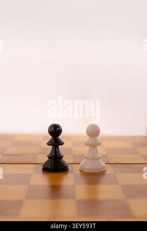 Eine vertikale Nahaufnahme eines einzelnen schwarzen Bauern und eines weißen Bauern, der von Angesicht zu Angesicht auf einem hölzernen Schachbrett steht. Der Hintergrund ist sauber, hell, Stockfoto