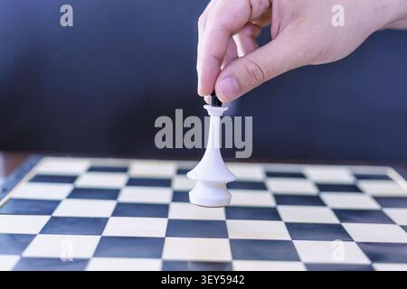 Nahaufnahme einer Hand, die einen weißen Schachkönig auf einem karierten Brett vor einem dunkelblauen Hintergrund platziert. Strategischer Wandel, Entscheidungsfindung und Führung Stockfoto