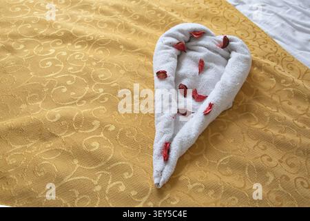 Herzförmige Handtuchdekoration mit Rosenblättern auf dem Hotelbett im Resortzimmer Stockfoto