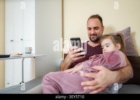Vater und Tochter entspannen sich zusammen im Bett und schauen auf den Smartphone-Bildschirm Stockfoto