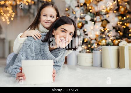 Brünette lächelnde weibliche Erwachsene und ihr schönes entzückendes kleines Kind in weißem Pullover im Wohnzimmer, Geschenk halten, Vater und Ehemann mit bevorstehendem Neujahr oder Weihnachten gratulieren Stockfoto