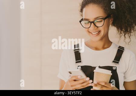 Entzückendes Mädchen mit lockigem Haar, überprüft Neuigkeiten, Nachrichten in sozialen Netzwerken, nutzt kostenlose Internetverbindung, trägt eine optische Brille, lässiges weißes T-Shirt und Overall, trinkt Kaffee zum Mitnehmen Stockfoto