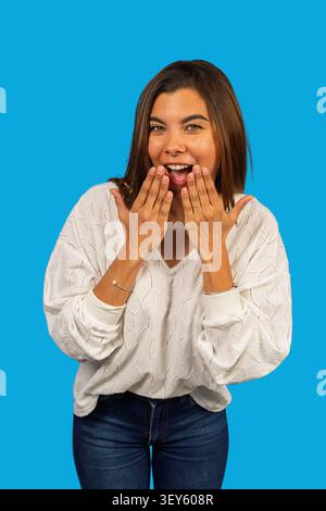 Junge Frau, die Überraschung und Freude ausdrückt, mit Händen, die den Mund auf blauem Hintergrund bedecken Stockfoto
