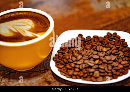 Eine Tasse Kaffee, dekoriert mit einem Herz und Kaffeebohnen auf einem weißen Teller. Dunkler Hintergrund Stockfoto