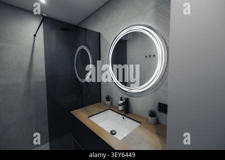 Modernes Bad mit schlankem Design und rundem Spiegel. Ein heller LED-Ring beleuchtet den Spiegelrahmen. Holzarbeitsfläche für Waschbecken und Minimalis Stockfoto