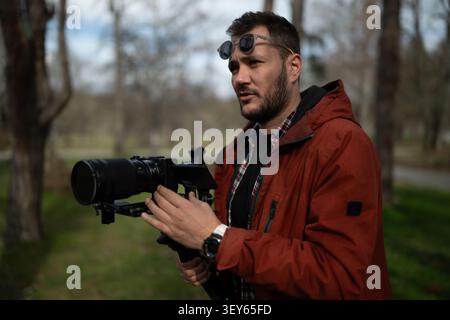 Ein Videofilmer in roter Jacke, der einen Kamerabildschirm bedient, während er im Freien im Park filmt Stockfoto