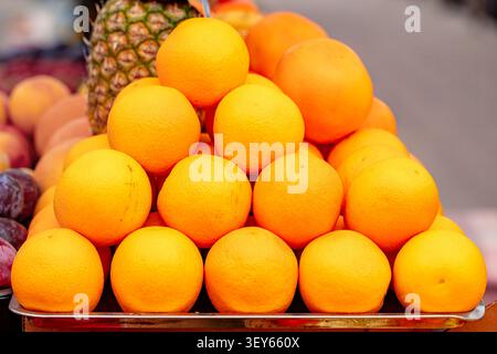Ein Anbieter stellt einen großen Haufen frischer Orangen auf einem Marktstand aus. In der Nähe sind andere Früchte wie Ananas und Pflaumen zu sehen. Die Szene zeigt ein geschäftiges Treiben Stockfoto