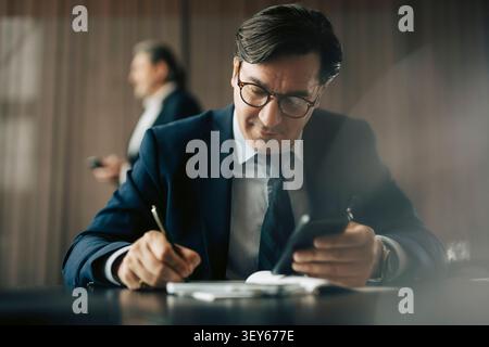 Reifer Geschäftsmann, der sein Smartphone hält, während er im Büro im Tagebuch schreibt Stockfoto