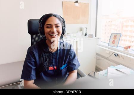 Porträt einer glücklichen weiblichen Gesundheitsberaterin, die ein Headset trägt, während sie auf dem Stuhl in ihrem Klinikbüro sitzt Stockfoto