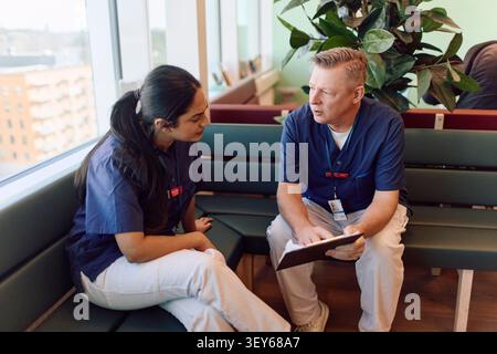 Zwei medizinische Fachkräfte diskutieren medizinische Dokumente, während sie auf der Bank im Wartezimmer des Krankenhauses sitzen Stockfoto