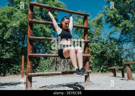 Junge Frau, die Outdoor-Fitnesstraining auf einem hölzernen Klettergerüst im sonnigen Park macht Stockfoto
