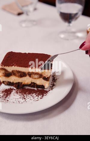 Tiramisu-Scheibe mit Kakaopulver auf weißer Platte, Gabel schneidet ein Stück. Stockfoto