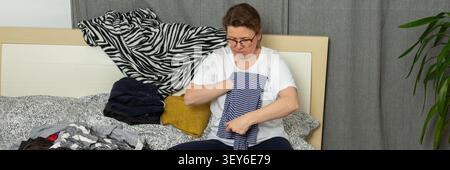Eine 52-jährige Frau sortiert Wäsche auf einem Bett. Sie hält ein Hemd und organisiert verschiedene herumliegende Kleidungsstücke. Ein Wäschekorb steht in der Nähe, während sie die anstehende Aufgabe erledigt, Banner Stockfoto