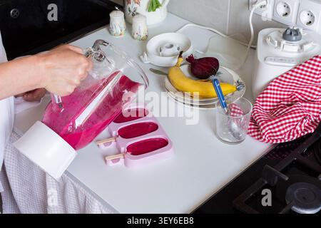 Person verwendet einen Mixer, um Erdbeeren, Bananen, Rüben und griechischen Joghurt in einer Küche zu vermischen. Eiscrememischung füllt Formen, während Küchenutensilien und Obst in der Nähe auf der Arbeitsplatte liegen. Stockfoto