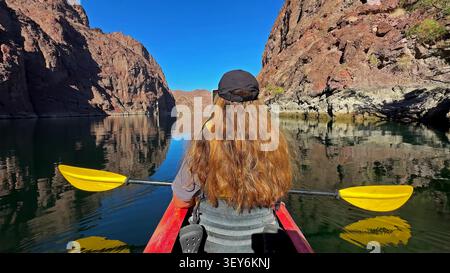 Kajakpaddeln Durch Den Red Rock Canyon. Rückansicht eines Kajakfahrers mit gelbem Paddel auf ruhigem Canyon-Wasser, umgeben von steilen roten Klippen, klarem Blau Stockfoto