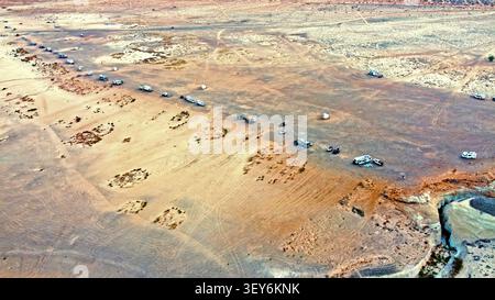 Vogelperspektive auf Wüstencampings und Offroad-Strecken. Luftaufnahme von Wohnmobilen, Autos und Lastwagen, die über Wüstenebenen mit staubigen Reifenspuren verstreut sind, schwach Stockfoto
