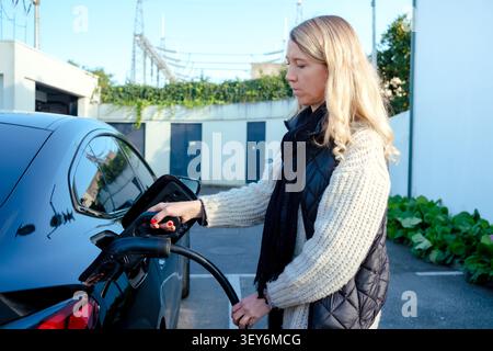 Frau, die ein elektrisches Autoladegerät an ihr Fahrzeug anschließt, fördert saubere Energie und umweltfreundliches Fahren Stockfoto