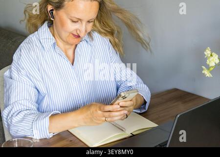 Frau mittleren Alters, die ihr Smartphone nutzt, nachdem sie am Arbeitsplatz einen kabellosen Ohrhörer aufgesetzt hat. Konzept für Sprachnachrichten, mobile Kommunikation, Musik-Streaming Stockfoto