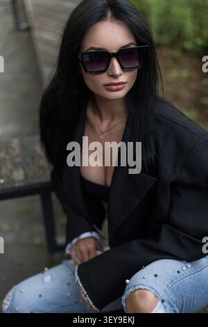Trendige junge Frau in übergroßem schwarzem Blazer, Sonnenbrille und verzierter, zerrissener Jeans sitzt draußen und blickt mit einer selbstbewussten, modernen Attitüde ab. Stockfoto
