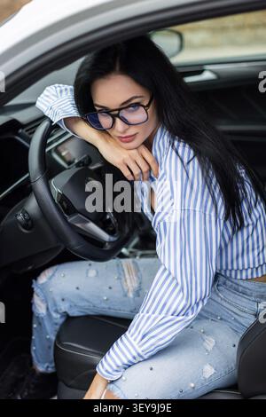 Junge Frau in Brille und gestreiftem Hemd posiert auf dem Fahrersitz, Hand auf Wange, lehnt sich am Lenkrad. Jeans mit Perlmuttmuster. Stockfoto