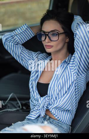 Eine schöne brünette Frau, die trendige Fliegerbrille trägt, entspannt sich in einem Autositz, fährt ihre Hände durch ihr Haar und blickt nachdenklich weg. Cas Stockfoto