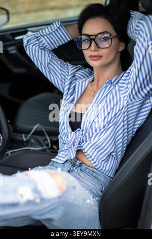 Attraktive junge Frau mit langen dunklen Haaren, die bequem in einer luxuriösen Fahrzeugkabine sitzt. Sie trägt lässigen Denim und eine gebundene gestreifte Bluse während des GA Stockfoto