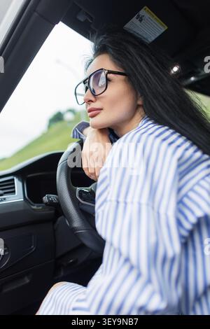 Trendige junge Fahrerin, die ihren Arm auf dem Lenkrad in einem modernen Fahrzeug-Cockpit ruht. Nachdenkliches Millennial-Porträt mit modischer Ausstattung Stockfoto