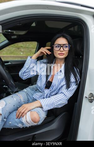 Eine attraktive Millennial-Frau in großer schwarzer Brille posiert auf dem Fahrersitz eines Autos und lehnt sich nach vorne. Lässiges Porträt des urbanen Lifestyles Stockfoto