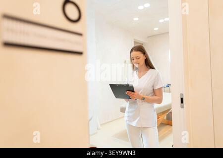 Eine junge Ärztin oder medizinische Assistentin steht in einem hellen Klinikraum, konzentriert auf das Schreiben auf einem Klemmbrett, betrachtet durch eine teilweise geöffnete Tür Stockfoto