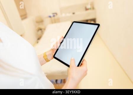 Hochwinkelaufnahme eines Mediziners oder Kosmetikers, der ein digitales Tablet mit leerem Bildschirmmockup in einem hellen, modernen Behandlungsraum der Klinik mit einem hält Stockfoto