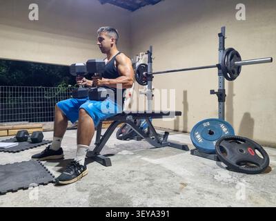 Muskulöser Mann, der mit schweren Kurzhanteln arbeitet, sitzt nachts auf der Hantelbank in der Garage. Bodybuilder-Training Kraft und Bizepskunst. Stockfoto