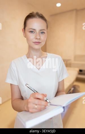 Nahaufnahme eines Porträts einer schönen jungen Ärztin oder Kosmetikerin, die in die Kamera schaut und mit einem Stift in ihrem Notizbuch gegen einen verschwommenen Mo schreibt Stockfoto