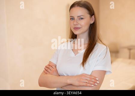 Professionelle Ärztin, Kosmetikerin oder Ärztin in weißer Uniform lächelnd mit gekreuzten Armen im Klinikinnenraum. Selbstbewusste junge Frauenphys Stockfoto