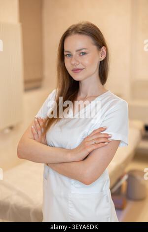Porträt einer schönen Ärztin oder Kosmetologin in weißer Uniform, die in die Kamera mit gekreuzten Armen schaut. Selbstbewusste Ärztin oder Ästhetik Stockfoto