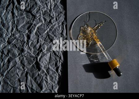 Tropfer, Der Mit Verschüttetem Gold Glitter Serum Auf Runder Glasschale Neben Zerknittertem Schwarzem Papier Auf Mattem Schwarzem Hintergrund Unter Angewinkelter Studio-Beleuchtung Liegt. Stockfoto