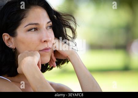 Nahaufnahme eines Porträts einer nachdenklichen jungen Frau, die ihr Kinn auf ihren Händen legt, während sie in einen grünen Park blickt. Natürliches Licht und Kopierspur Stockfoto