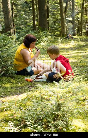 Eine Frau und ein Junge sitzen auf dem moosigen Boden in einem Wald und teilen sich eine Mahlzeit mit gekochten Würstchen. Sie wirken entspannt und glücklich und genießen ihre Outdoor-Aktivitäten Stockfoto